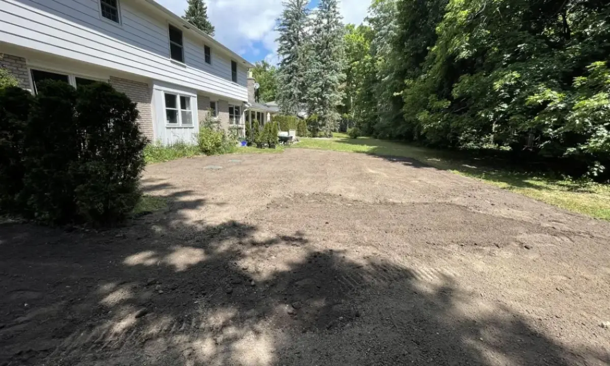 A large residential backyard in Hamilton after complete old sod removal, showing freshly graded bare dirt next to a two-story home and mature trees, prepared for new turf.